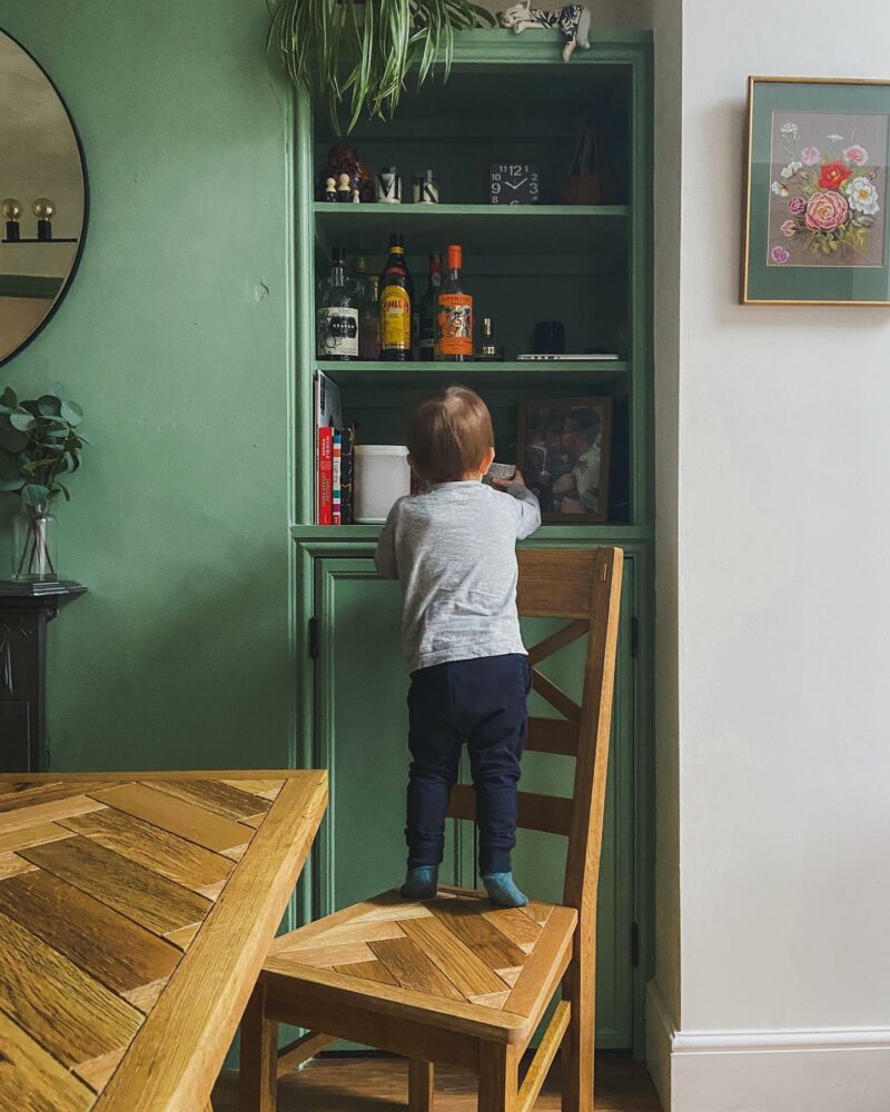 Vibrant green dining room with a Parquet dining table and a little boy reaching up to the in-built shelves.