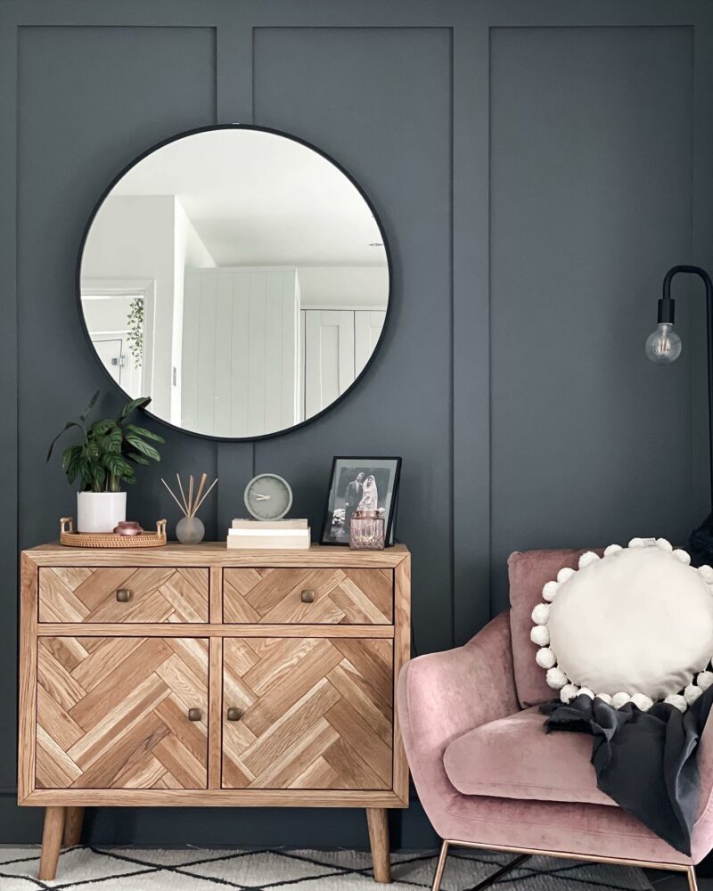 Cosy living room nook with dark grey panelled walls, a pink velvet chair and a small Oak Furnitureland Parquet sideboard topped with plans and decorative objects.