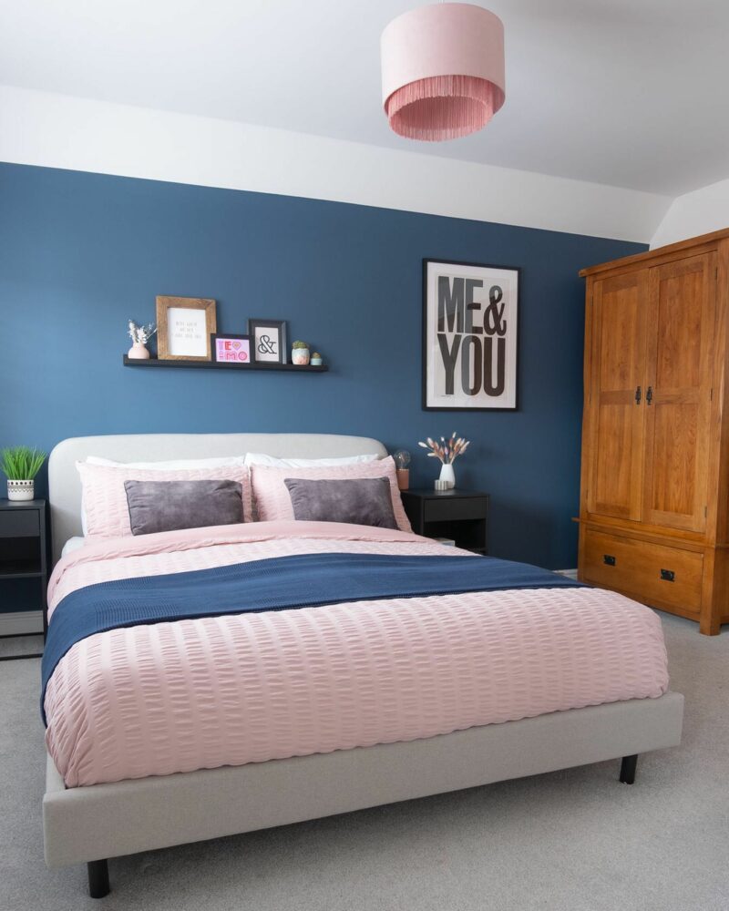Bedroom with dark blue walls, grey upholstered bed, Original Rustic oak wardrobe, and pink bedding and lighting. 
