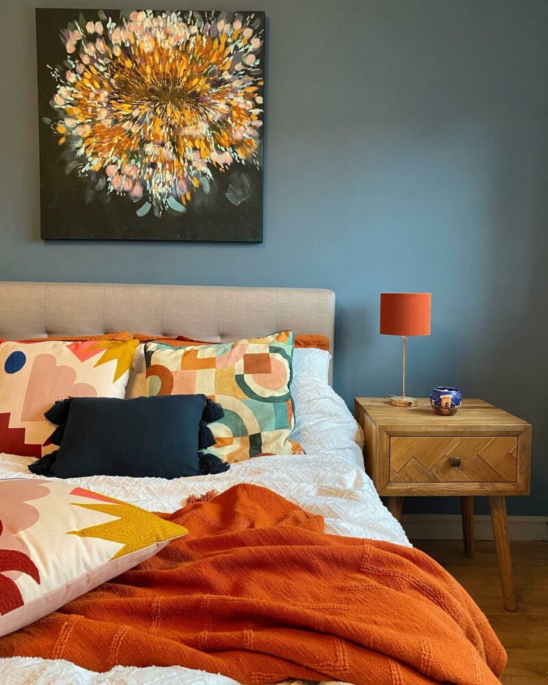 Colourful bedroom with dark walls, statement art, bright cushions and bedding, and a Parquet bedside table.