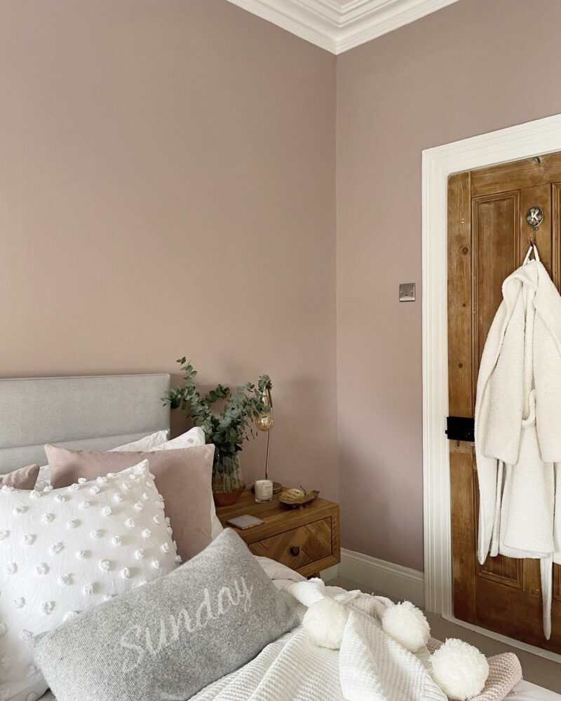 Close up of a bed filled with white and pink cushions and a Parquet bedside table, with light pink bedroom walls adding a contrast. 
