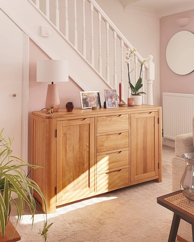 Pale pink entrance hall featuring an Oak Furnitureland Romsey sideboard topped with a lamp, photos, plants and candles.