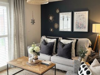 A sofa-living room furniture-beige sofa-houndstooth armchair-charcoal feature wall-monochrome cushions-wood and metal coffee table-wicker lampshade-floor standing lamp-jute rug-neutral linen curtain-wooden flooring-abstract wall art