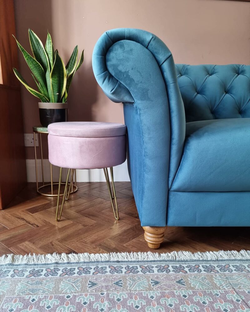 Blue velvet Oak Furnitureland Montgomery Chesterfield-style sofa wtih pink side table, plant and patterned rug.