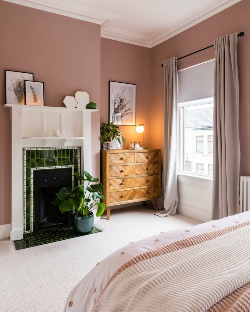 Salmon pink bedroom featuring Parquet chest of drawers, fireplace with green tiles, and house plants.