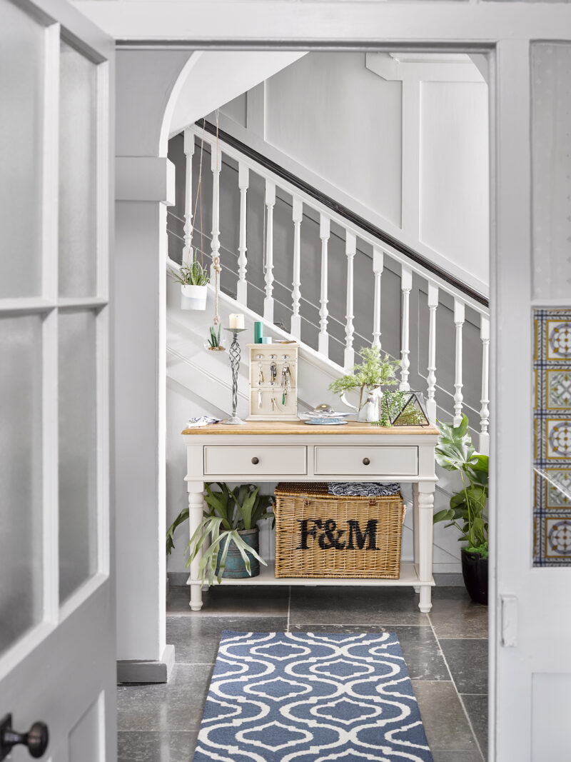 Oak Furnitureland Shay almond grey console table, placed in a hallway and styled with plants and decorative accessories.
