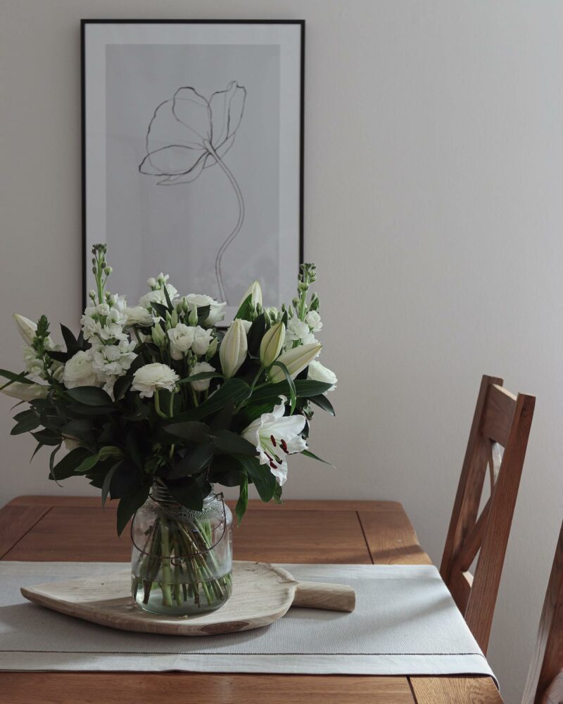 Oak dining table with a vase of beautiful white flowers and simple art behind.