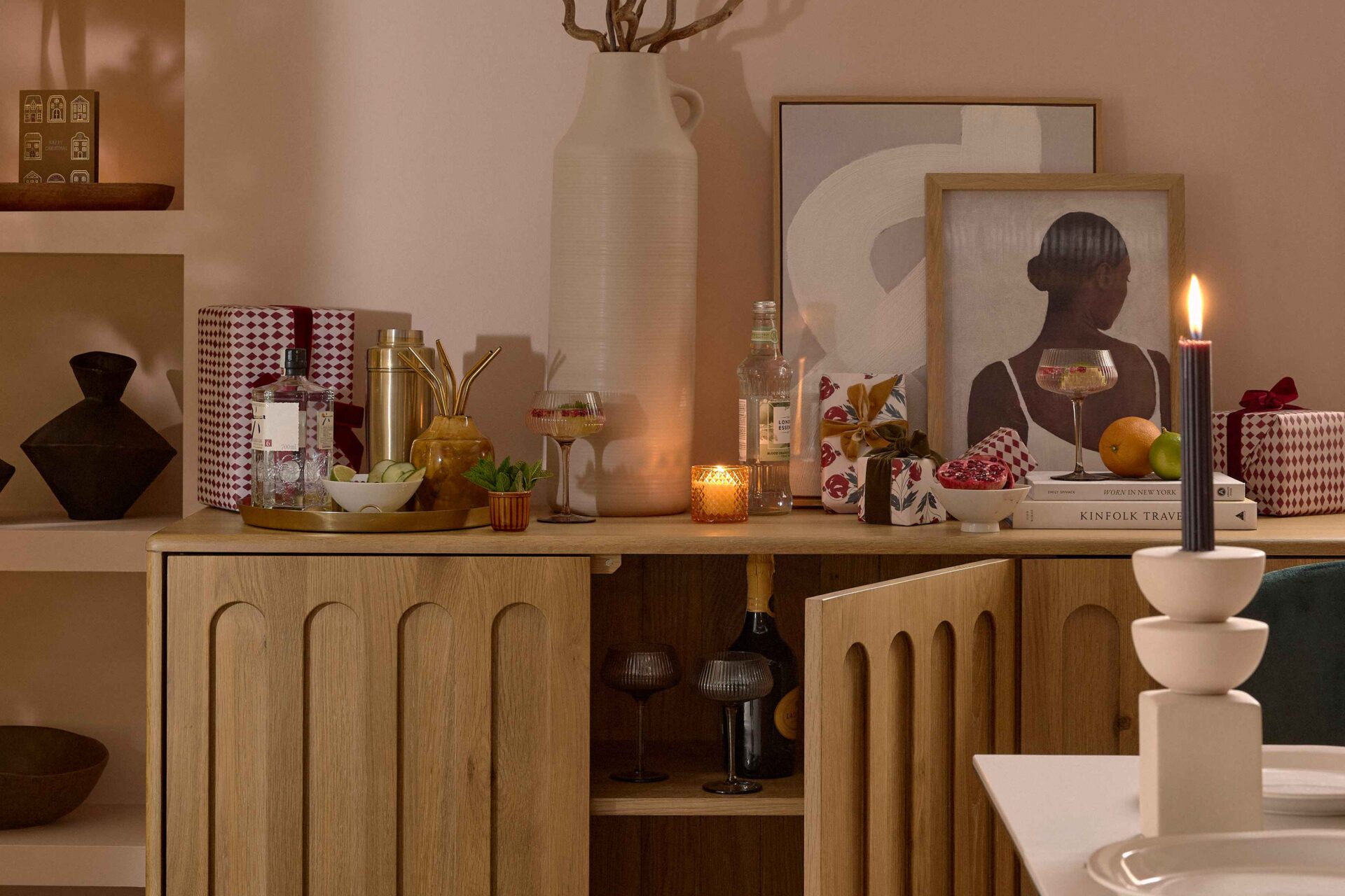 A sideboard-dining room furniture-arch detail wooden sideboard-abstract artwork-pink blush walls-cocktail shaker-wrapped presents-metallic tray-spirits-mixers-straws-cocktail glasses-fresh mint and cucumber-oversized cream vase-sculptural candle holder-lit candles-built in shelving