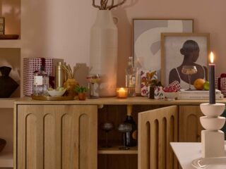 A sideboard-dining room furniture-arch detail wooden sideboard-abstract artwork-pink blush walls-cocktail shaker-wrapped presents-metallic tray-spirits-mixers-straws-cocktail glasses-fresh mint and cucumber-oversized cream vase-sculptural candle holder-lit candles-built in shelving