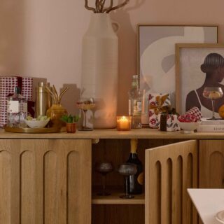 A sideboard-dining room furniture-arch detail wooden sideboard-abstract artwork-pink blush walls-cocktail shaker-wrapped presents-metallic tray-spirits-mixers-straws-cocktail glasses-fresh mint and cucumber-oversized cream vase-sculptural candle holder-lit candles-built in shelving