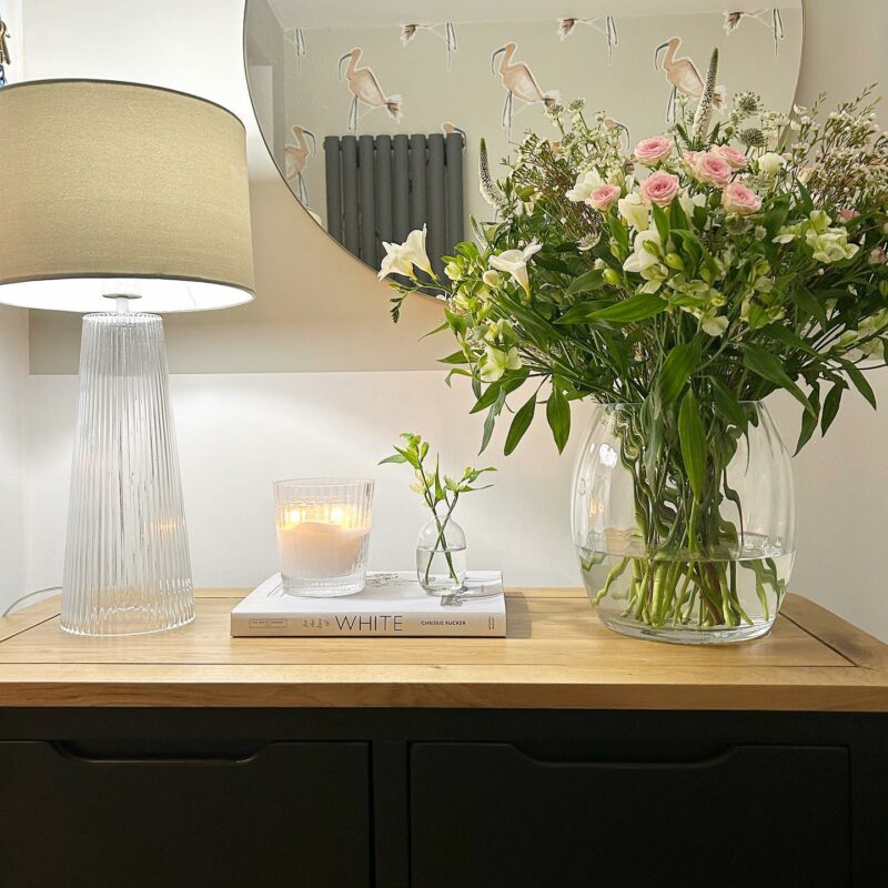 Oak Furnitureland slate grey Grove console table close up with a glass lamp and a bouquet of flowers.
