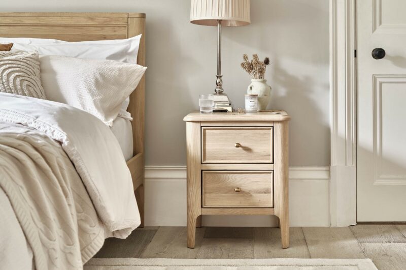 Close up shot of the Oak Furnitureland Newton natural oak bedside table with a lamp and vase on top, next to a bed with neutral bedding and cushions.
