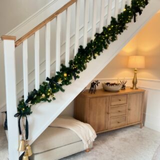 A staircase and sideboard-hallway furniture-natural oak large sideboard-hallway storage-neutral fabric footstool-beige carpet-white staircase-festive garland-brass table lamp