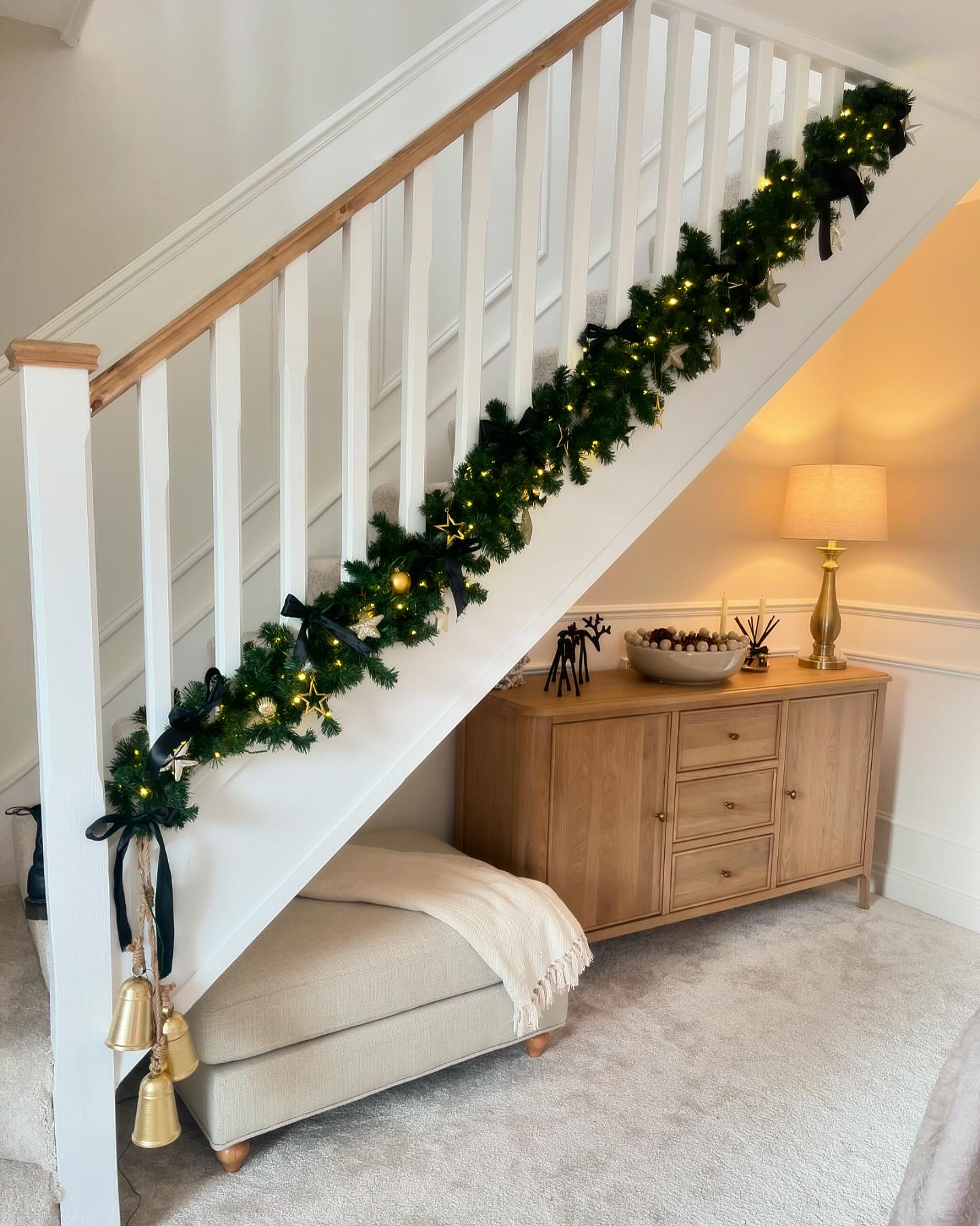 A staircase and sideboard-hallway furniture-natural oak large sideboard-hallway storage-neutral fabric footstool-beige carpet-white staircase-festive garland-brass table lamp