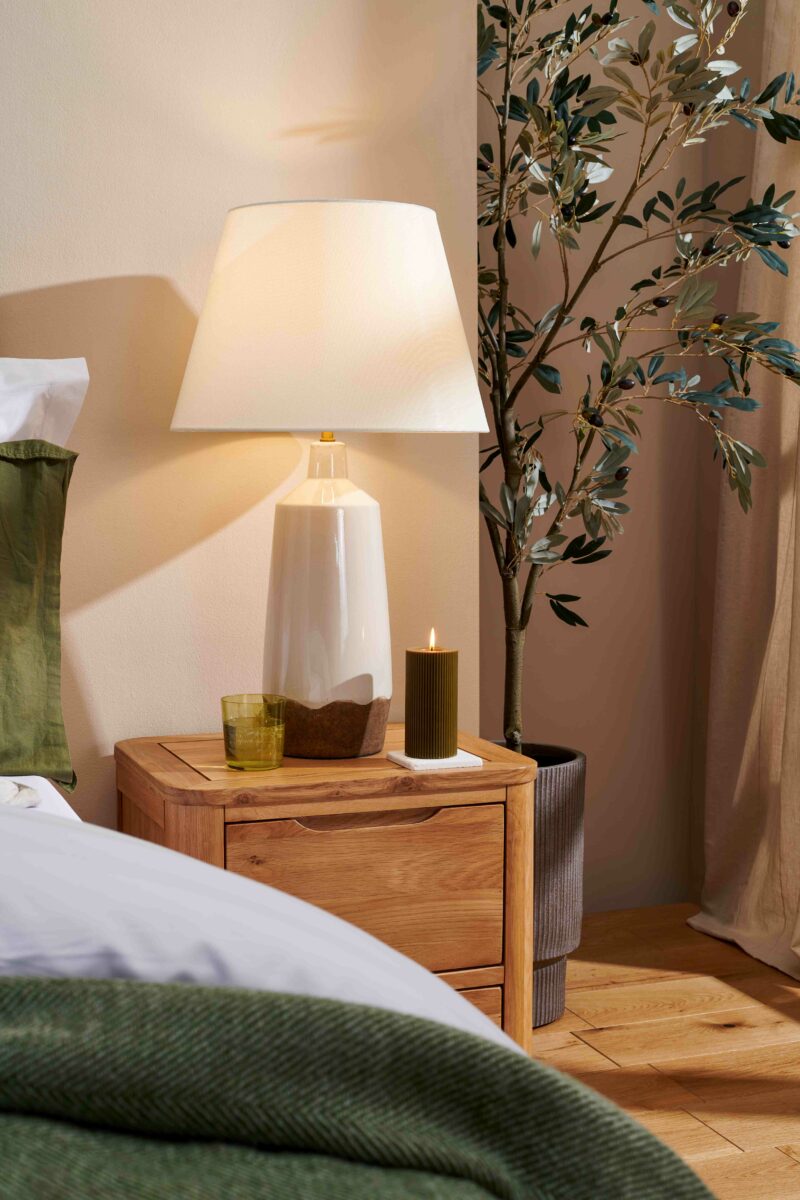 Close up of an oak bedside table with a cream lamp on top, next to a bed with white bedding and a dark green throw.