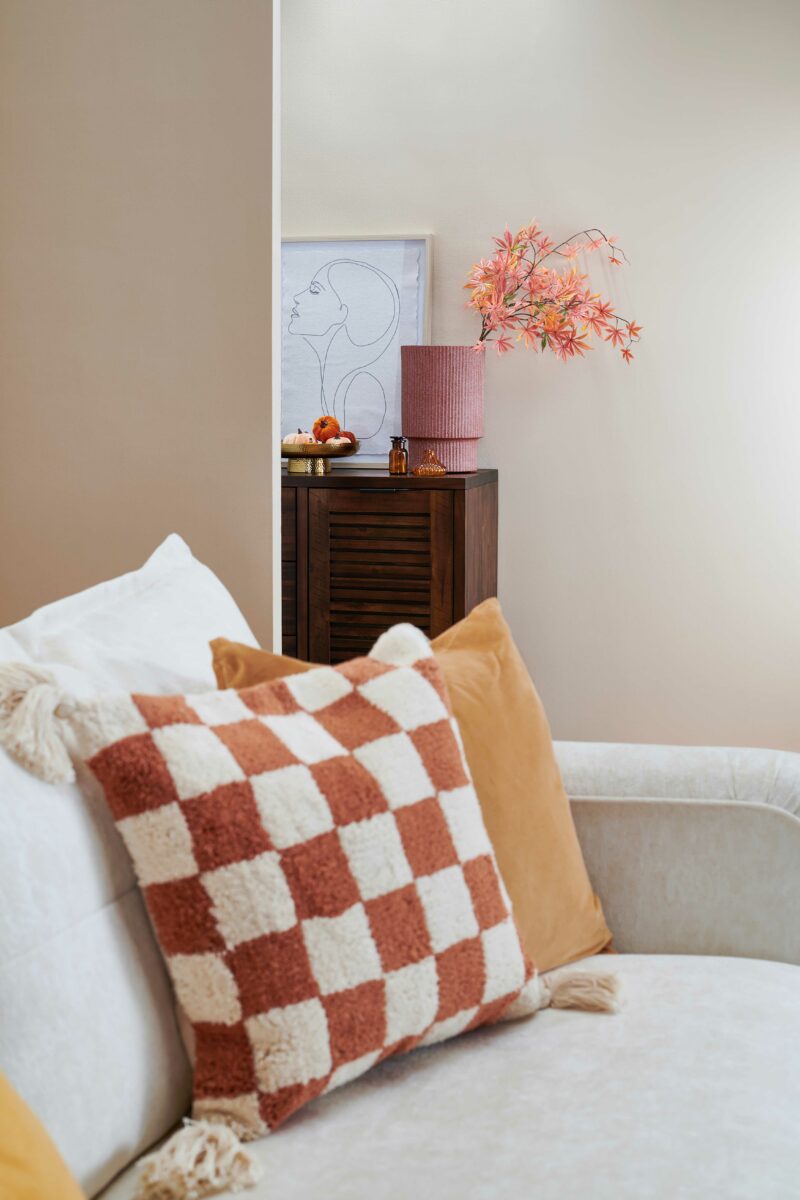 Close up of a cream sofa with orange and cream checkered cushions and a dark -stained oak sideboard in the background.