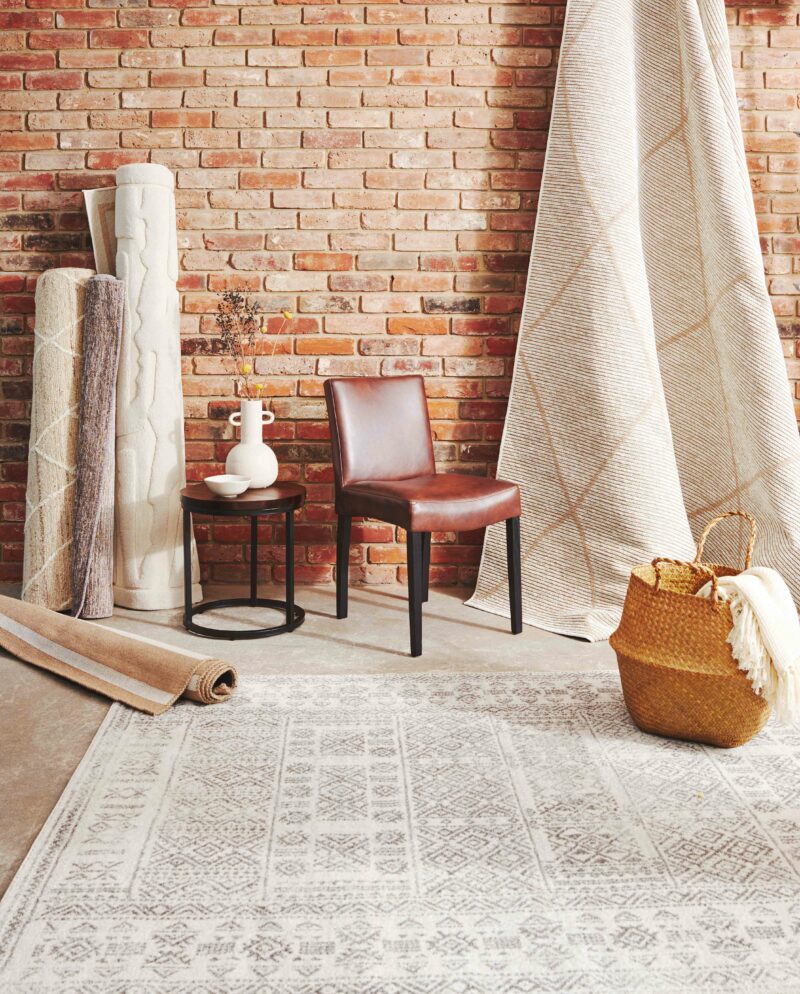 Brick wall with cream rolled up rugs, a brown leather chair and a dark acacia and metal side table, with a cream rug in the foreground.