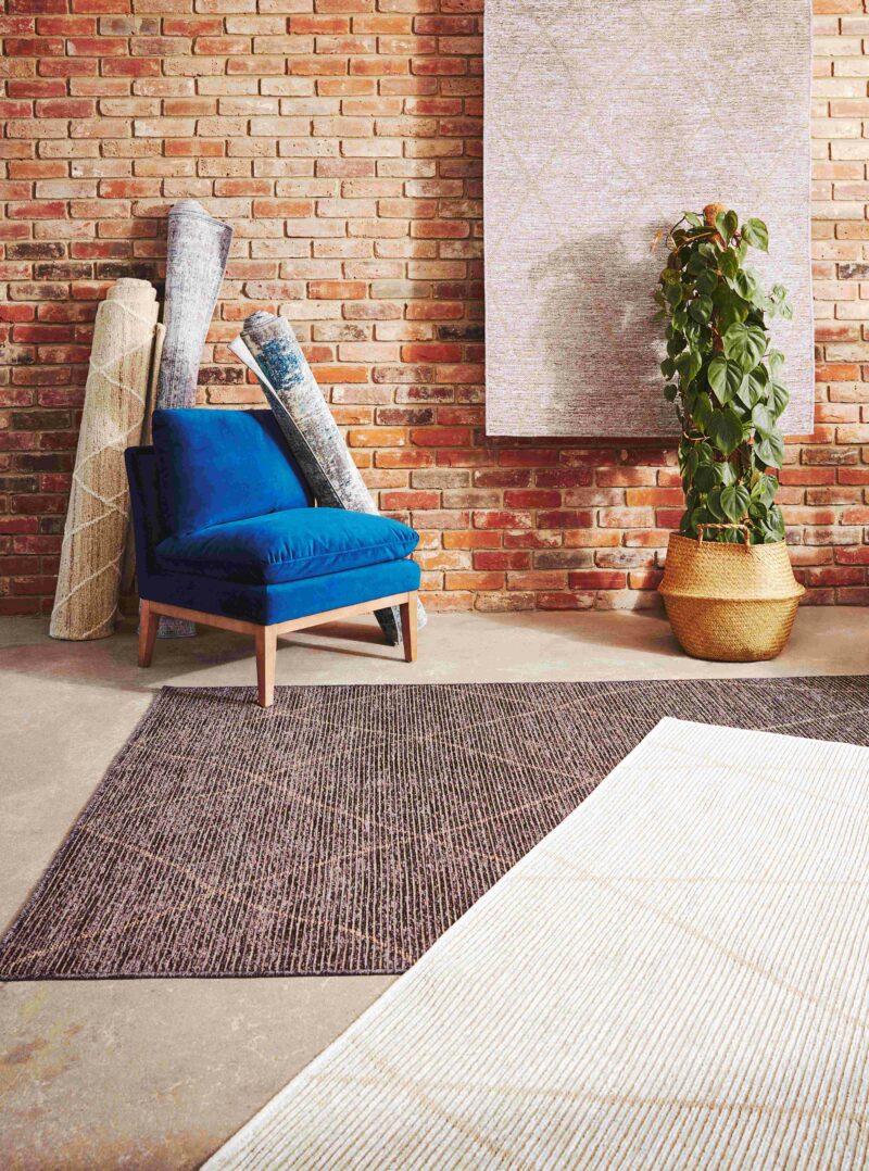 Vibrant blue velvet accent chair with three rugs rolled up against a brick wall, with a large plant also in shot.