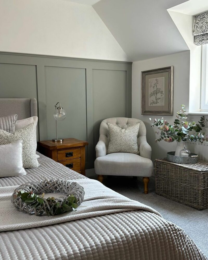 Country farmhouse-style bedroom with sage green wall panelling, a cream chair, a close-up of the bed and a rustic oak bedside table.
