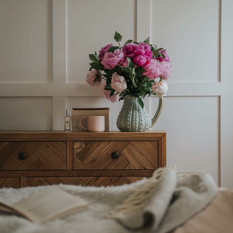 Close up of Parquet chest of drawers with a jug of pink peonies, perfume, and books on top, wtih a bed in the foreground.