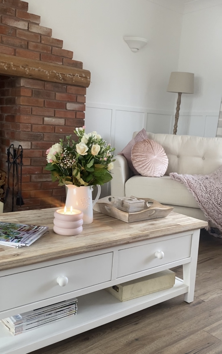 Oak Furnitureland cream loveseat styled with pink cushions and a throw, featuring a table in the foreground with flowers and a candle on.