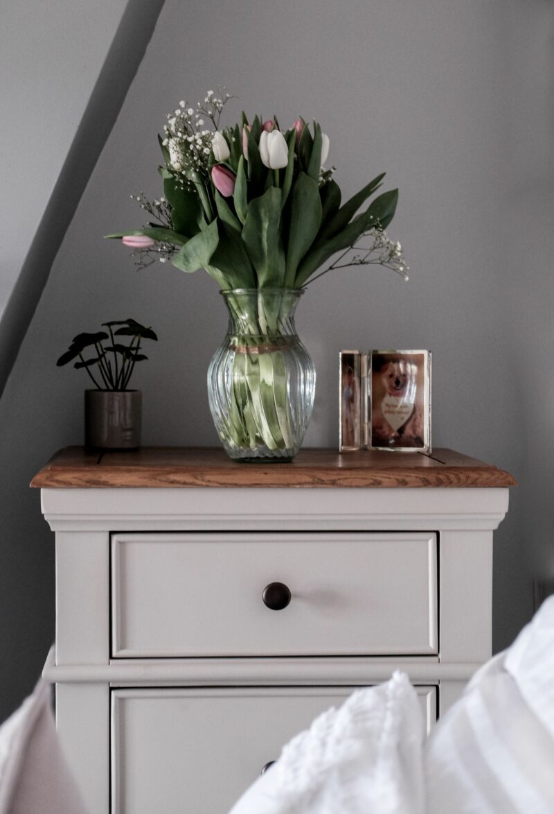 Oak Furnitureland grey and oak tallboy chest, topped with a vase of tulips, plant & photo frame. 