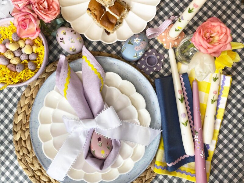 Easter tablescape flat lay on a gingham tablecloth with pastel napkins, candles and painted eggs.