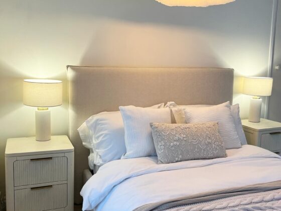 A bed and bedside table-bedroom furniture-white walls-white bedding-neutral knitted throw-Art Deco style bedside table-upholstered bed-bedside table lamps-cloud ceiling light-neutral carpet