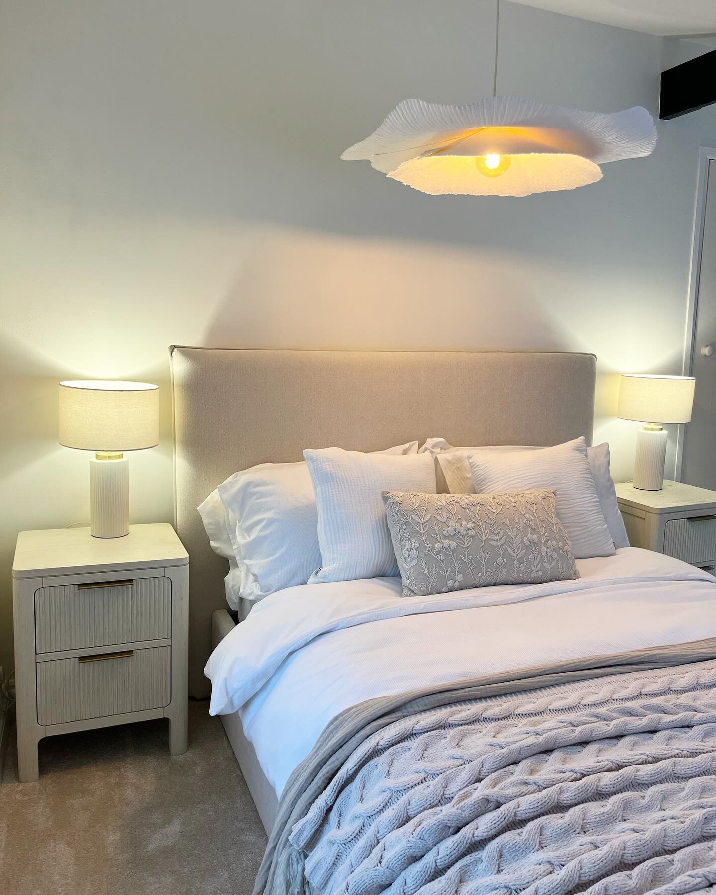 A bed and bedside table-bedroom furniture-white walls-white bedding-neutral knitted throw-Art Deco style bedside table-upholstered bed-bedside table lamps-cloud ceiling light-neutral carpet