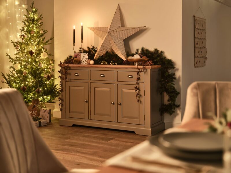 Grey painted sideboard styled for Christmas with a large star and foliage and a Christmas tree in the background.