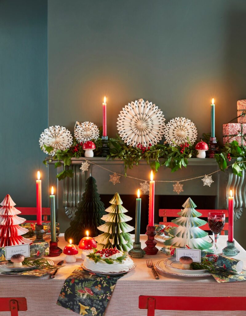 Red, white, and green Christmas tablescape, with a decorated fireplace behind including paper decorations and candles.
