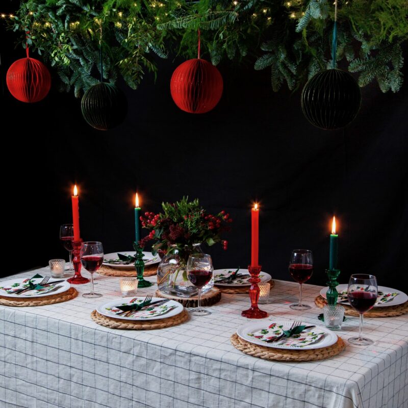 Table styled for Christmas with a check tablecloth, red and green candles, with greenery and red and green honeycomb decorations hanging above.