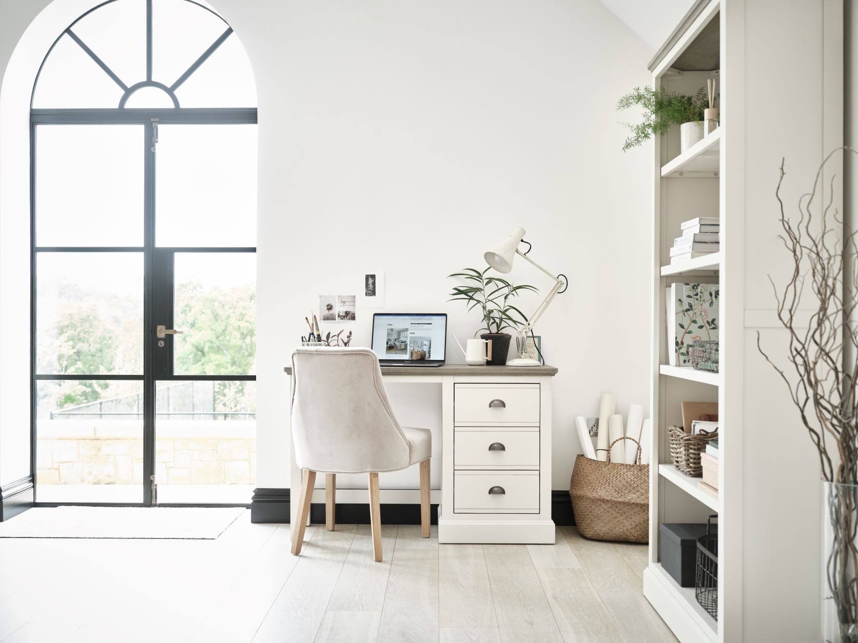 White painted desk paired with a grey velvet chair in a light and airy white home office with a large door out to the garden.