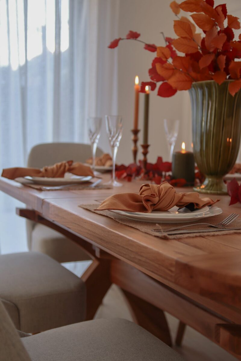 A close up of an Oak Furnitureland Hercules dining table with light grey Bette dining tables around it. The table is set for dinner with jute placemats, white plates, cutlery, orange napkins, a leafy table garland, wine glasses, candles and a green vase filled with orange autumnal foliage.