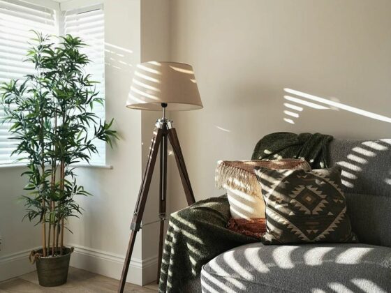 Sunlight spills through slatted blinds into a living room. A large houseplant and floor lamp stand next to a grey Oak Furnitureland Jensen sofa that has been styled with a cosy throw and scatter cushions.