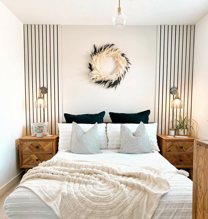 Two Oak Furnitureland Parquet bedside tables sit either side of a double bed. The bed has been made with decorative pillows and a cream blanket. A black and cream pampas grass wreath hangs on the wall above the bed.