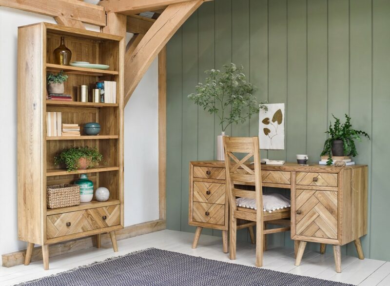 Oak Furnitureland Parquet bookcase styled with decorative accessories in a home office. A Parquet desk and wooden chair sit adjacent to the bookcase against a green panelled wall.