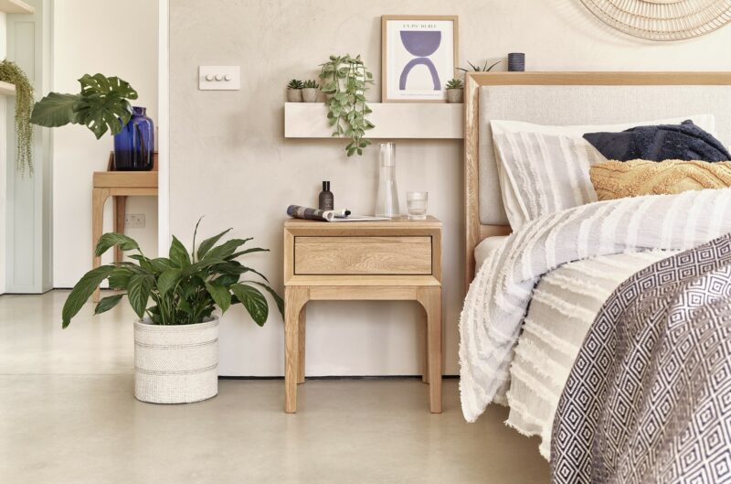 Oak Furnitureland Aston bedside table and oak and upholstered bed, in a bedroom with houseplants and wall art.