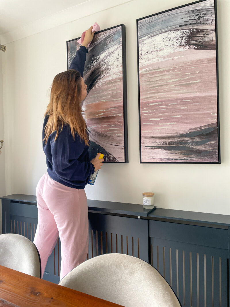 Woman cdusting an Oak Furnitureland pink and grey piece of artwork in dining room above a radiator.