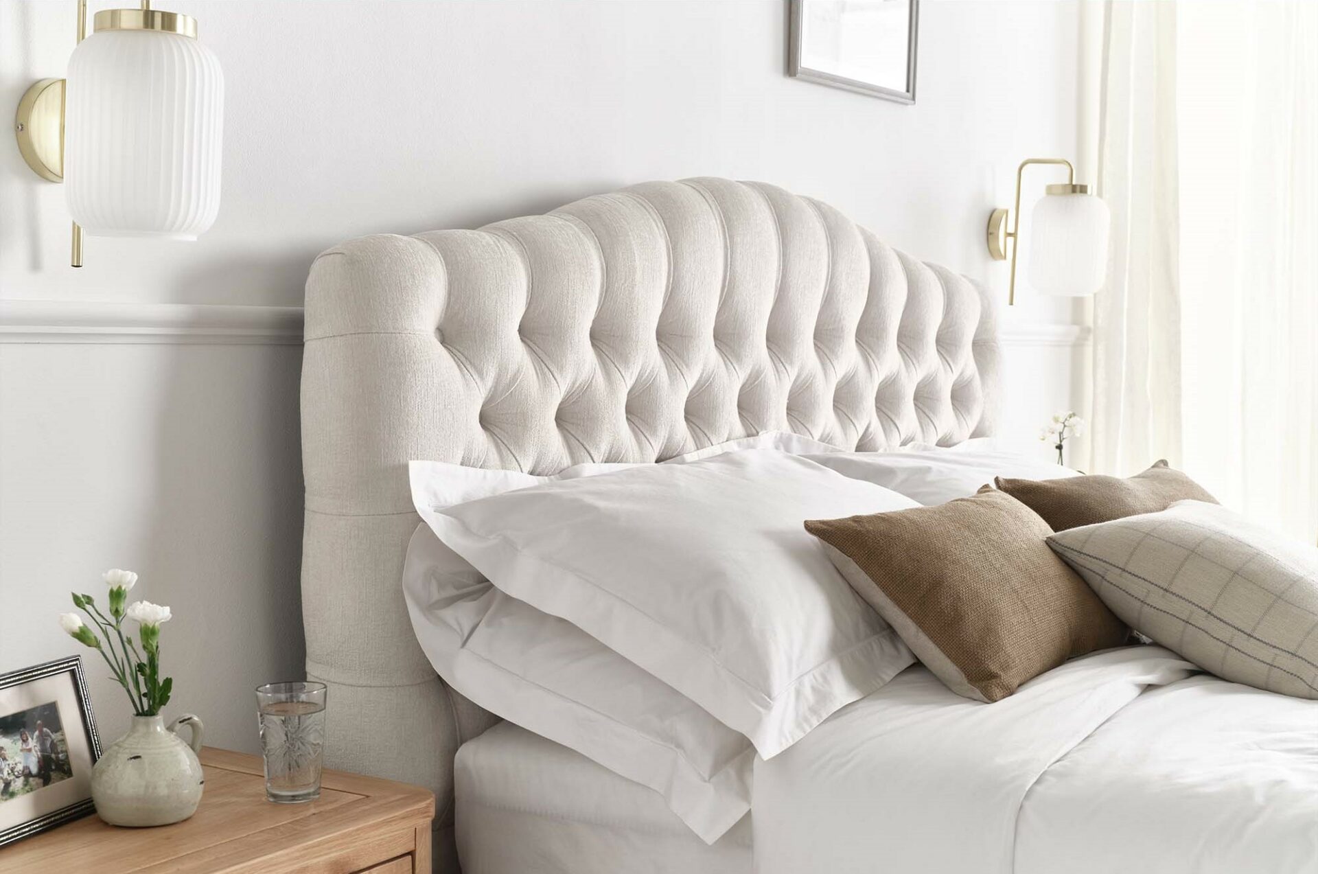 Pale grey Chesterfield-style upholstered bed with brown cushions and gold and white bedside wall lights.