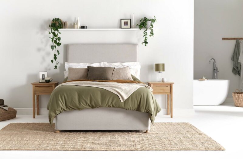 Grey upholstered bed with olivve green bedding, neutral cushions and Scandi-style bedside tables, with en-suite in the background.
