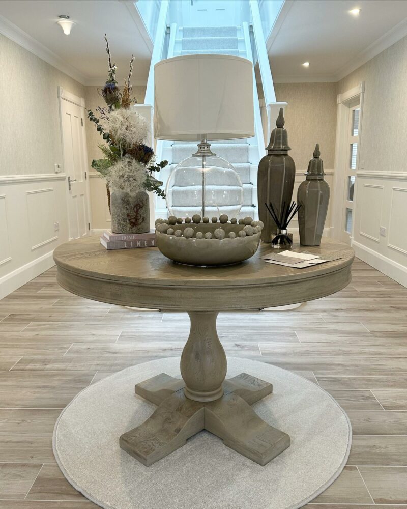 Large washed oak pedestal round table toped with decorative accessories in a neutral hallway.