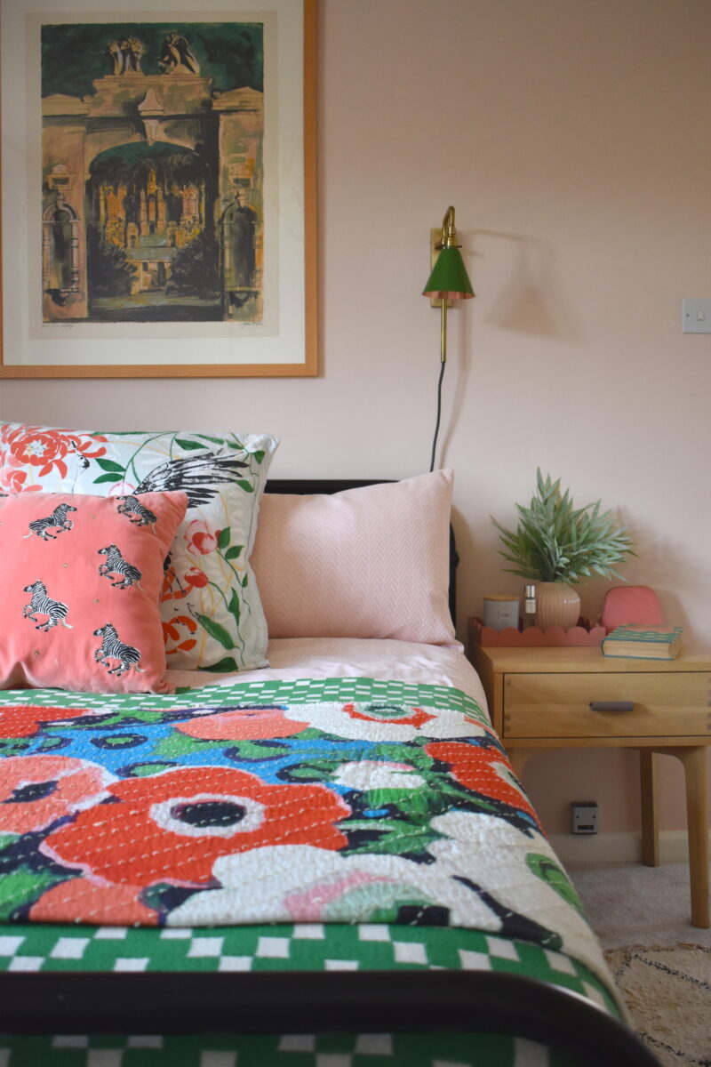 Close up of a brightly coloured bed, wtih artwork overhead, a wall light and bedside table topped with a plant.