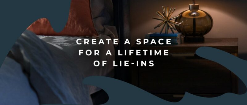 Dark lit bedroom with a close up of a bed and a bedside table with a lamp on. 