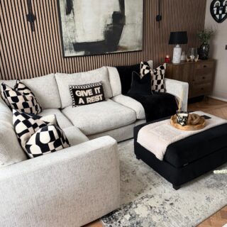 A sofa, footstool, and sideboard-living room furniture-beige corner modular sofa-black footstool-black and white geometric cushions-black throw-neutral abstract rug-wooden floor-wooden panelling on feature wall-large abstract monochrome painting-black wall lights-beige wall-dark wood sideboard-wooden scallop tray-cream throw
