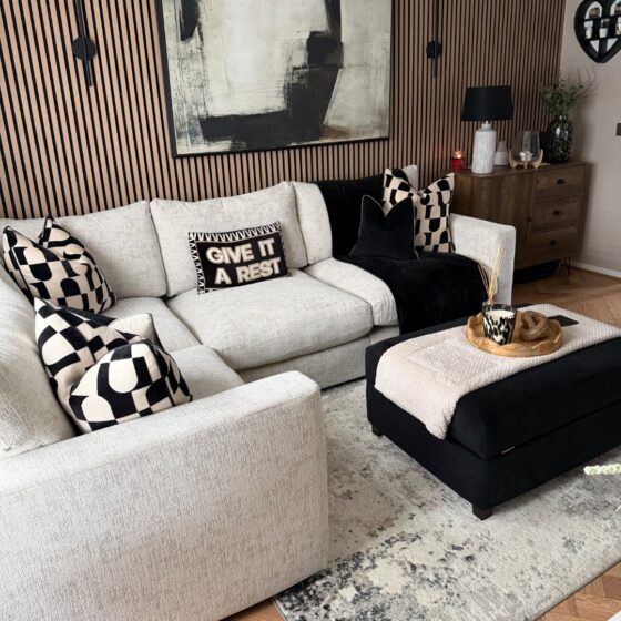 A sofa, footstool, and sideboard-living room furniture-beige corner modular sofa-black footstool-black and white geometric cushions-black throw-neutral abstract rug-wooden floor-wooden panelling on feature wall-large abstract monochrome painting-black wall lights-beige wall-dark wood sideboard-wooden scallop tray-cream throw