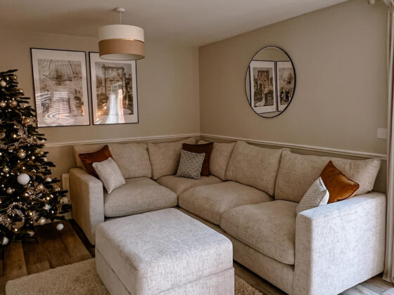 A sofa and Christmas tree-living room furniture-textured neutral rug-cream walls-cream corner sofa-storage footstool-Christmas tree with gold decorations-round wall mirror
