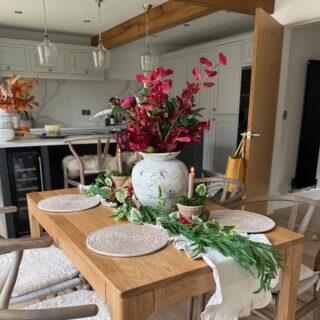 A dining table-dining room furniture-natural oak wooden table-Christmas table decor-festive foliage-pine garland runner-woven placemats-wishbone dining chairs-open plan kitchen diner-exposed wooden beams-grey kitchen units-neutral walls