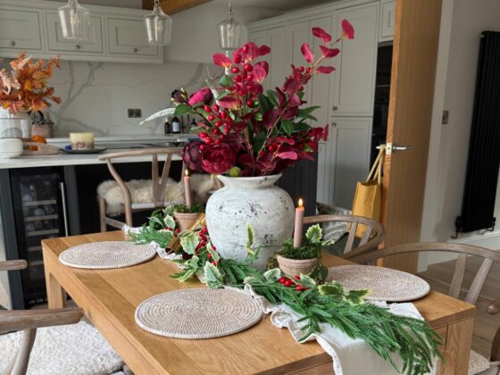A dining table-dining room furniture-natural oak wooden table-Christmas table decor-festive foliage-pine garland runner-woven placemats-wishbone dining chairs-open plan kitchen diner-exposed wooden beams-grey kitchen units-neutral walls
