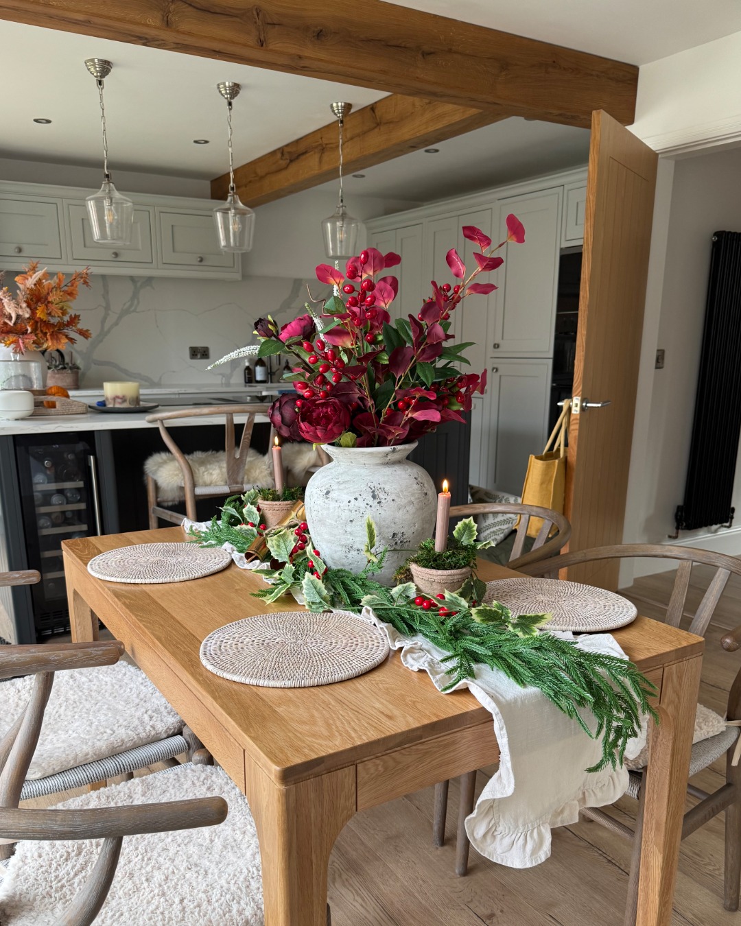 A dining table-dining room furniture-natural oak wooden table-Christmas table decor-festive foliage-pine garland runner-woven placemats-wishbone dining chairs-open plan kitchen diner-exposed wooden beams-grey kitchen units-neutral walls